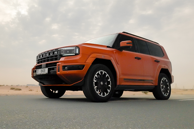 Front three-quarter profile of a matte orange Jetour rugged SUV parked on an asphalt desert roadside.