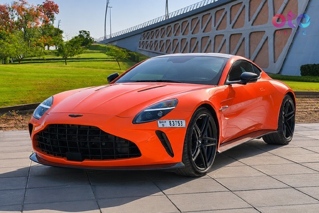 Glossy orange Aston Martin luxury sports coupe car parked on a stone walkway under a modern concrete bridge.